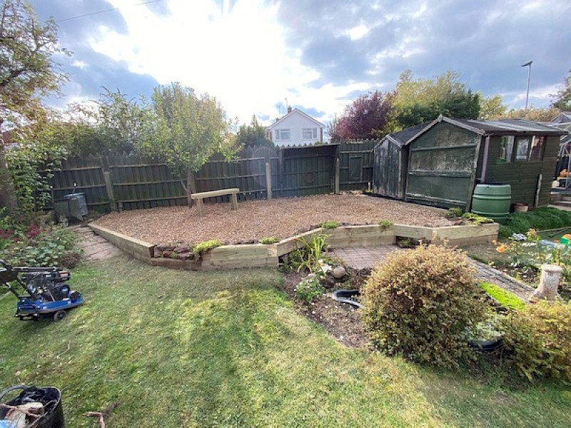 Backyard garden with gravel area, wooden retaining walls, a small bench, and surrounding plants, showcasing landscaping and gardening services