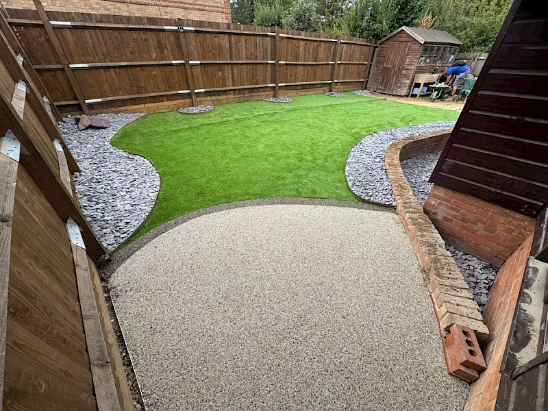 Resin bound patio in a landscaped back garden, creating a neat, low-maintenance seating area beside a wooden shed, lawn and mature shrubs.
