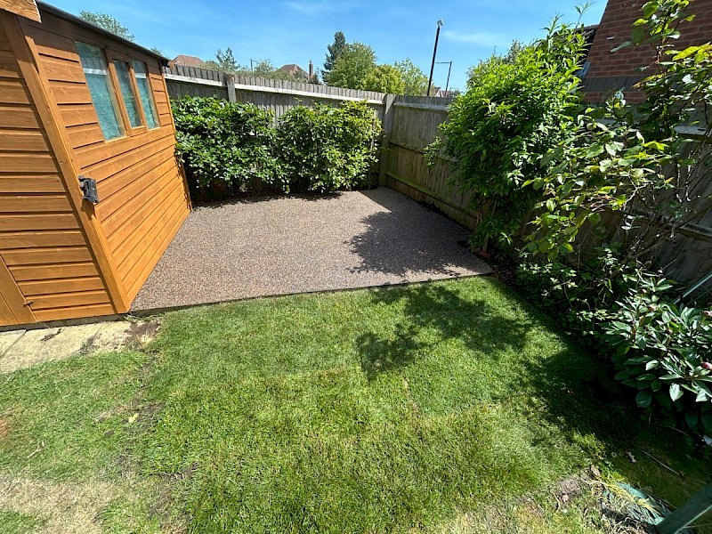 Resin bound patio in a landscaped back garden, creating a neat, low-maintenance seating area beside a wooden shed, lawn and mature shrubs.